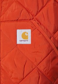 Orange quiltat tyg med diagonala sömmar; har en vit rektangulär etikett med en gul logotyp och "Carhartt" i lila text.