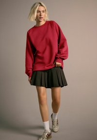 Punainen ylisuuri collegepaita yhdistetty mustaan taittopäähineeseen. Mallilla on valkoiset sukat ja hopeiset lenkkarit, joissa on viininpunaisia yksityiskohtia. Pehmeä rakenne.