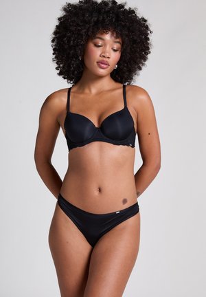 Frau mit lockigem Haar, die einen schwarzen BH und passende schwarze Unterwäsche trägt, steht mit den Händen hinter dem Rücken vor einem einfarbigen Hintergrund.