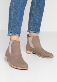 Gray suede ankle boots with elastic side panels and a low stacked wooden heel. Worn with blue denim jeans, showcasing a casual style.