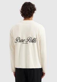 Long-sleeve cream shirt featuring a black printed text design on the back that reads "Pure Path - Own the Journey." Soft fabric, relaxed fit.