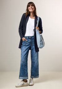 Blazer lungo blu navy, t-shirt bianca, jeans a gamba larga azzurri, e sneakers bianche. Tiene una borsa a tracolla azzurra. Outfit informale.