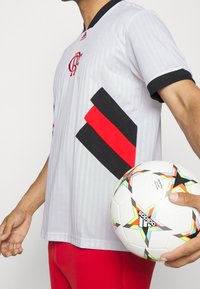 Maillot de football gris avec des rayures diagonales noires et rouges, col noir, manches courtes, et logo adidas, associé à un ballon de football blanc avec des accents colorés.