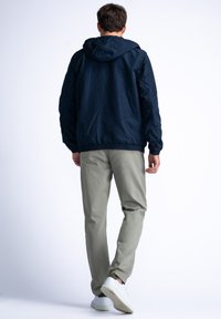 Tmavě modrá mikina s kapucí a elastickými manžetami a spodním lemem, v kombinaci se světle šedými kalhotami a bílými teniskami, zachycena zezadu.