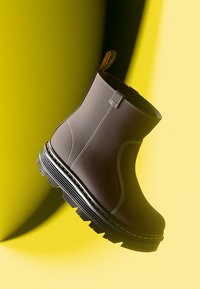 Botín marrón de tobillo con material similar al cuero, costuras blancas, suela negra gruesa y un acento amarillo alrededor de la base; cuenta con una lengüeta de tirón.