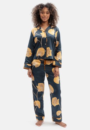 Woman with curly hair wearing dark blue satin pajamas with large yellow flower prints, standing barefoot against a plain background.