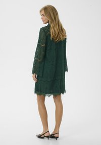Krajková šaty v tmavě zelené barvě, ke kolenům, s dlouhými rukávy, s vroubkovaným lemem a složitými vzory. Nosit s černými botami na nízkém podpatku.