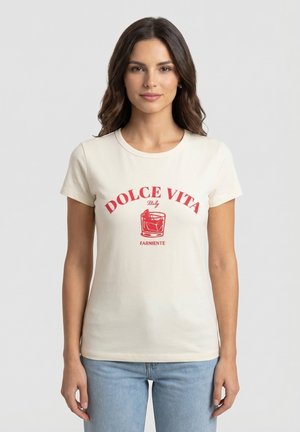 Mujer con cabello largo y castaño que lleva una camiseta blanca con texto rojo "DOLCE VITA Italy" y un gráfico de copa de cóctel, combinada con jeans azul claro.