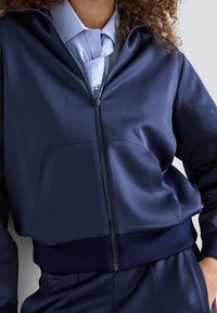 Tmavě modrá bunda na zip z hladkého materiálu, s vysokým límcem, přední kapsou a žebrovanými manžetami a lemem.