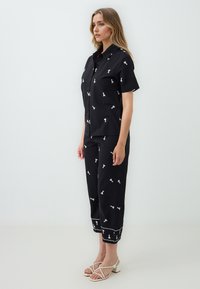 Czarna koszulka z krótkim rękawem i spodnie w stylu cropped z białymi haftowanymi wzorami. Spodnie mają wykończenie z białymi akcentami. Modelka nosi sandały na paski.