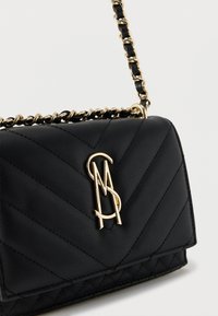 Μαύρο τσόκερ από δερματίνη με χρυσαφί αλυσίδα και κούμπωμα με χρυσό λογότυπο Steve Madden σε απλό ανοιχτό φόντο.