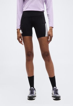 Persona vistiendo pantalones cortos deportivos negros con bolsillos laterales y rayas blancas, calcetines negros, zapatillas grises y una camiseta de manga larga lila.