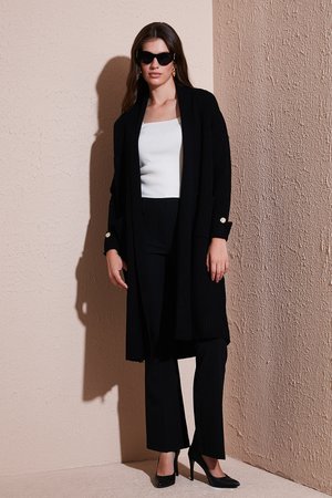Black ribbed long cardigan with side pockets, worn over a white top and black trousers. Black high heels complement the outfit.