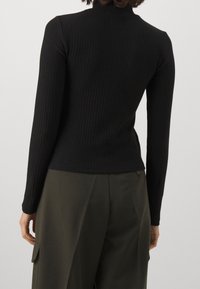 Svart ribbad långärmad topp med hög halsringning. Materialet är stretchigt, åtsittande och slätt. Kombineras med olivgröna vida byxor.