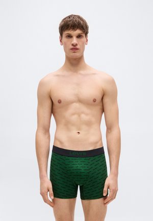 Νέος άνδρας με κοντά μαλλιά που φοράει σκούρα πράσινα μπόξερ Levi's με μαύρη μέση και λογότυπο, στέκεται μπροστά σε απλό φωτεινό φόντο.