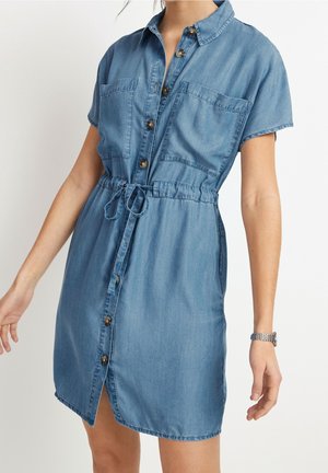 Femme portant une robe en denim bleu à manches courtes avec boutons sur le devant, deux poches poitrine et un cordon à la taille, debout devant un fond blanc.
