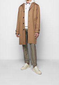 Hnědý kabát s přiléhavým střihem, velkým límcem a předními knoflíky přes šedou mikinu. V kombinaci s béžovými kalhotami a krémovými teniskami.