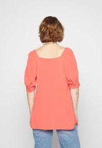 Top in corallo con maniche a sbuffo realizzato in tessuto leggero. Presenta una scollatura quadrata e una vestibilità morbida, con una texture sottile e una finitura liscia.