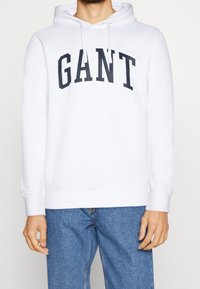 Sudadera blanca con "GANT" impreso en azul marino en el pecho. Presenta una capucha con cordón y puños acanalados, combinada con unos vaqueros de color azul claro.