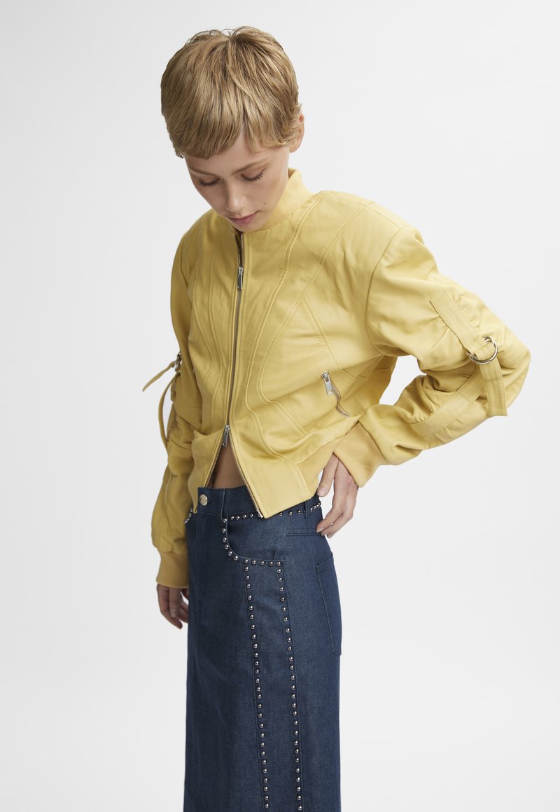 Veste bomber en cuir jaune avec poches zippées et couture texturée, associée à une jupe en denim taille haute ornée d'accentuations en clous.