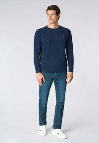 Maglione girocollo a maglia blu navy con polsini a costine, caratterizzato da un piccolo logo rosso sul petto, abbinato a jeans blu e sneakers bianche.