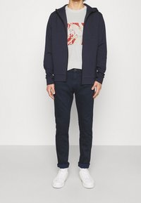 Námořnická mikina na zip s tkaničkami, světle šedé tričko s potiskem, tmavě modré džíny a bílé tenisky. Hladká tkanina, casual střih.