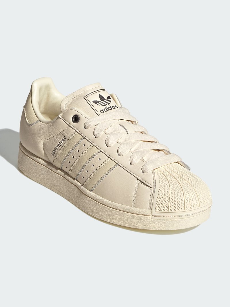 Μπεζ αθλητικό παπούτσι Adidas Superstar με προστασία δακτύλων (shell toe), τρεις χαρακτηριστικές ρίγες, κείμενο "Superstar" και λογότυπο Adidas στη γλώσσα, απεικονισμένο σε κλίση προς τα αριστερά.