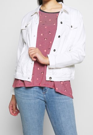 Personne portant un jean bleu clair, un haut rayé rouge et noir avec de petits motifs floraux, et une veste blanche déboutonnée.