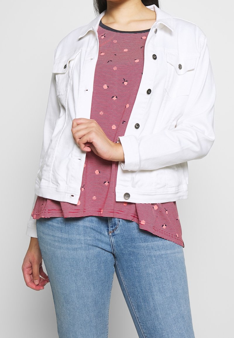 Personne portant un jean bleu clair, un haut rayé rouge et noir avec de petits motifs floraux, et une veste blanche déboutonnée.