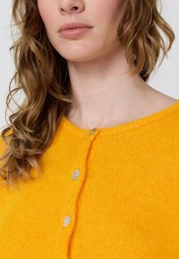 Geltonas megztas kardiganas su apvalia iškirpte, trimis šviesios spalvos sagomis ir minkšta, tekstūruota medžiaga.