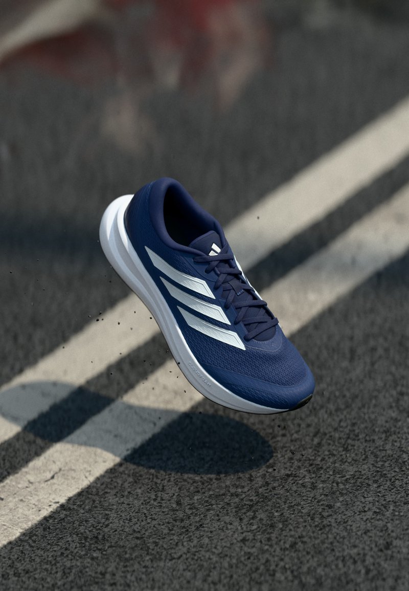 Chaussure de course Adidas bleue avec des bandes blanches et une semelle rembourrée, suspendue au-dessus d'une route en asphalt avec des marquages blancs au sol.