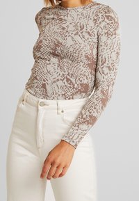 Langärmliges, tailliertes Oberteil aus strukturiertem Beige mit einem braunen Schlangenmuster, kombiniert mit weißen, hochgeschnittenen Jeans.