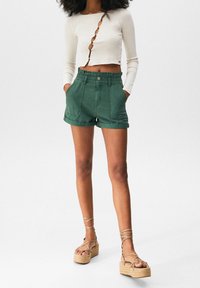 Mujer con un top corto blanco de manga larga con cortes diagonales, pantalones cortos verdes de talle alto y sandalias de plataforma beige con tiras en el tobillo.