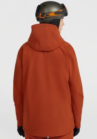 Oranje skijas met een capuchon, voorzien van een gladde, matte textuur. De achterkant toont een subtiele logo. Gecombineerd met zwarte handschoenen.