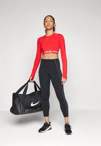 Crvena dugih rukava crop majica s "NIKPRO" brendingom, u kombinaciji s crnim tajicama koje imaju bijele pruge. Crna Nike torba za teretanu upotpunjuje izgled.