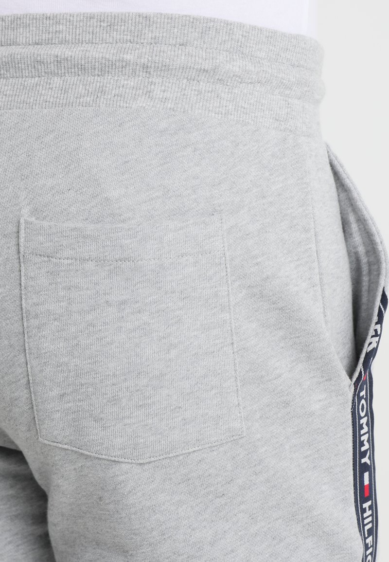 Pantalons de jogging en coton gris avec une poche arrière et une bande latérale ornée du logo de la marque dans des couleurs bleu marine, rouge et blanc. Texture lisse.