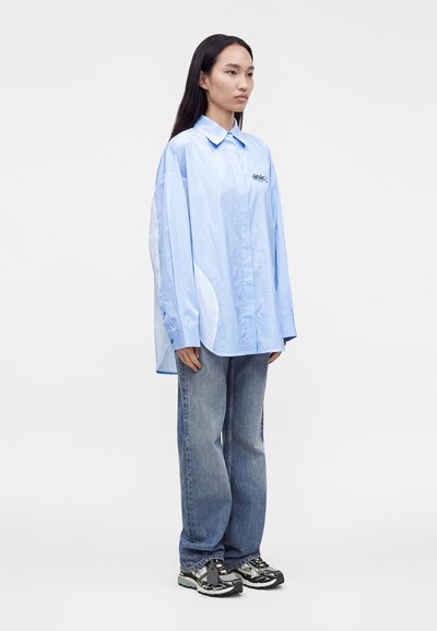 Femme aux longs cheveux foncés portant une chemise oversize rayée bleu clair, un jean large bleu et des baskets noires et blanches, debout devant un fond uni.