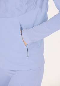 Ljusblå jacka med en strukturerad övre del, ficka med dragkedja och ribbade ärmar. Tillverkad av mjukt, lätt tyg, perfekt för aktiv användning.