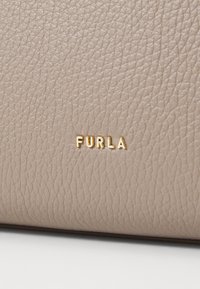 Beige leather handbag with a textured surface, featuring a gold "FURLA" logo accent. The design is minimalist and elegant.