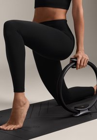 Fekete legging és crop top, amit egy személy visel, fekete Pilates gyűrűt tartva, térdelve egy textúrált fekete jóga szőnyegen.