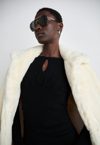Black fitted dress with a keyhole neckline and pleated detail, paired with a cream faux fur coat. Dark sunglasses and hoop earrings complete the look.