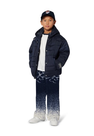 Doudoune marine avec fermeture éclair, t-shirt blanc, pantalons dégradés bleu-blanc à motifs, et baskets blanches. Le chapeau présente un design graphique.