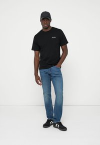 T-shirt preta com logotipo bordado, jeans de ganga azul e ténis pretos com riscas brancas. Os acessórios incluem uma capa preta.