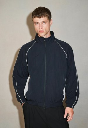 Young man with short curly hair wearing a navy blue track jacket with white piping, standing against a plain light background.