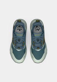 Sinised ja hallid maastikujooksujalatsid tekstureeritud pealsega, väljapaistva paelumise süsteemiga ning kollaste rõhkudega keelel ja küljel.