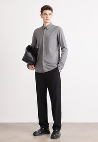 Grijze lange mouwen overhemd met knopen en een kraag, gecombineerd met zwarte broeken en schoenen. Het model houdt een zwarte leren tas vast.