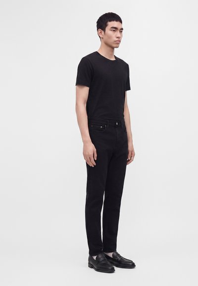 Jeune homme debout, portant un t-shirt noir ajusté, un pantalon noir et des mocassins noirs, sur un fond gris clair uni.