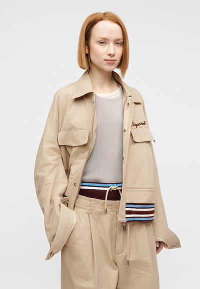 Μπεζ βαμβακερό μπουφάν με δύο μπροστινές τσέπες, σε συνδυασμό με ανοιχτό γκρι ριμπ τοπ και μπεζ παντελόνια με πολύχρωμη ριγέ ζώνη μέσης.