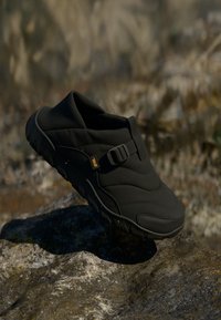 Pantofii negri slip-on pentru exterior, cu o talpă robustă și o curea ajustabilă, așezati pe o suprafață stâncoasă, luminată de soare, în aer liber.