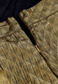 Gepolstertes Metallicgewebe in Gold mit einem Diamantmuster, das einen Reißverschlussdetail aufweist. Der Hintergrund ist ein dunkler Stoff, möglicherweise schwarz.